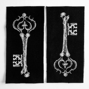 May include: Two black fabric patches with white skeleton key designs. The keys have ornate details and are in a vintage style.
