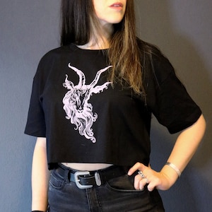 以下が含まれることがあります： 角のあるヤギの頭がピンクのグラフィックプリントされた、黒いクロップドTシャツです。