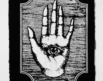 Puede incluir: Parche bordado en blanco y negro de una mano con un ojo en la palma, enmarcado en un borde decorativo.