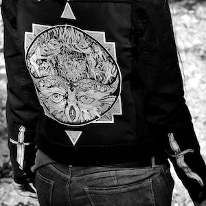Op de afbeelding: Zwart denim jack met een witte grafische print van een uil met een derde oog, omringd door een geometrisch ontwerp. Het jack heeft een dolkdesign op elke mouw.