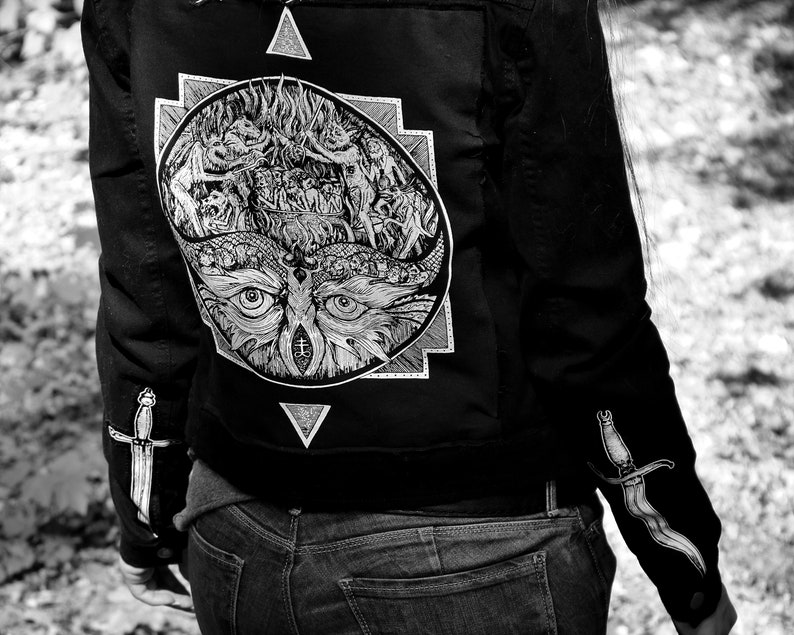 Op de afbeelding: Zwart denim jack met een witte grafische print van een uil met een derde oog, omringd door een geometrisch ontwerp. Het jack heeft een dolkdesign op elke mouw.