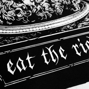 Puede incluir: Parche blanco y negro con una imagen estilizada de una cabeza de le&oacute;n y el texto "eat the rich" en una fuente g&oacute;tica.
