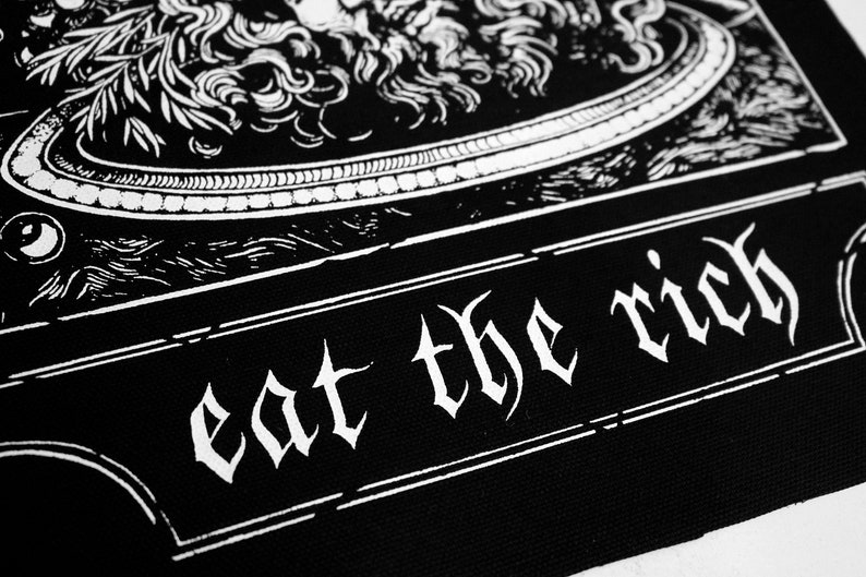 Puede incluir: Parche blanco y negro con una imagen estilizada de una cabeza de le&oacute;n y el texto "eat the rich" en una fuente g&oacute;tica.