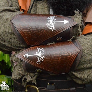 Könnte beinhalten: Ein Paar braune Lederarmschützer mit einem silbernen Baumdesign. Die Schützer werden an den Unterarmen getragen und haben eine spitze Form.