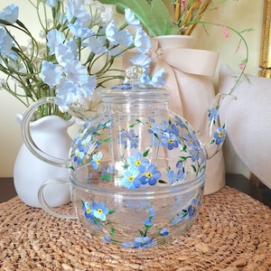 May include: A clear glass teapot and matching teacup stackable set featuring hand-painted blue forget-me-not flowers with yellow centres and green stems. The teapot includes a glass lid with a small knob handle and a curved spout, while the teacup has a side handle.