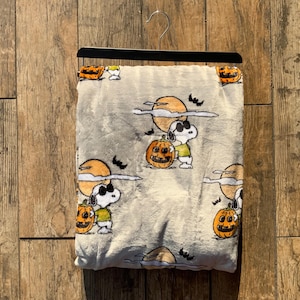 May include: A light beige plush throw blanket featuring a repeating pattern of Snoopy wearing sunglasses, orange jack-o'-lantern pumpkins, yellow full moons, and small black bats. The blanket is displayed hanging from a black clothes hanger.