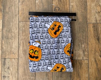 May include: A white fabric garment featuring a repeating pattern of black-outlined jack-o'-lanterns and orange pumpkins. Some pumpkins contain a small white cartoon dog character. The fabric is folded over a black plastic hanger with a metal hook.