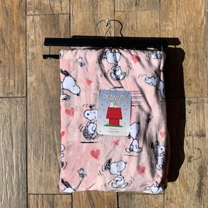 May include: A light pink throw blanket featuring a repeating pattern of Snoopy in various poses and small red hearts. A rectangular product tag is attached to the centre, displaying the text PEANUTS and an illustration of a red doghouse.