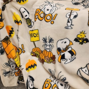 May include: A cream-coloured Halloween-themed blanket featuring Snoopy and Woodstock characters. The blanket has orange pumpkins, the word "BOO!" in orange, and Snoopy dressed in various Halloween costumes. The blanket is made of a soft, plush material.