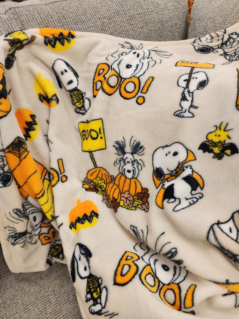 May include: A cream-coloured Halloween-themed blanket featuring Snoopy and Woodstock characters. The blanket has orange pumpkins, the word "BOO!" in orange, and Snoopy dressed in various Halloween costumes. The blanket is made of a soft, plush material.