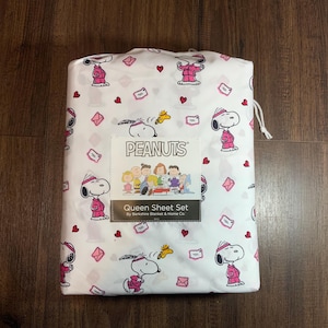 Puede incluir: Juego de sábanas Queen blancas con Snoopy con atuendo rosa, corazones y sobres. El juego incluye la palabra "PEANUTS" y un gráfico de la pandilla Peanuts. El embalaje está atado con un cordón blanco.