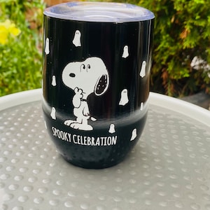 May include: Black stainless steel tumbler with a clear lid. The tumbler features a white illustration of Snoopy and ghosts, with the words "Spooky Celebration". The tumbler is designed for Halloween.