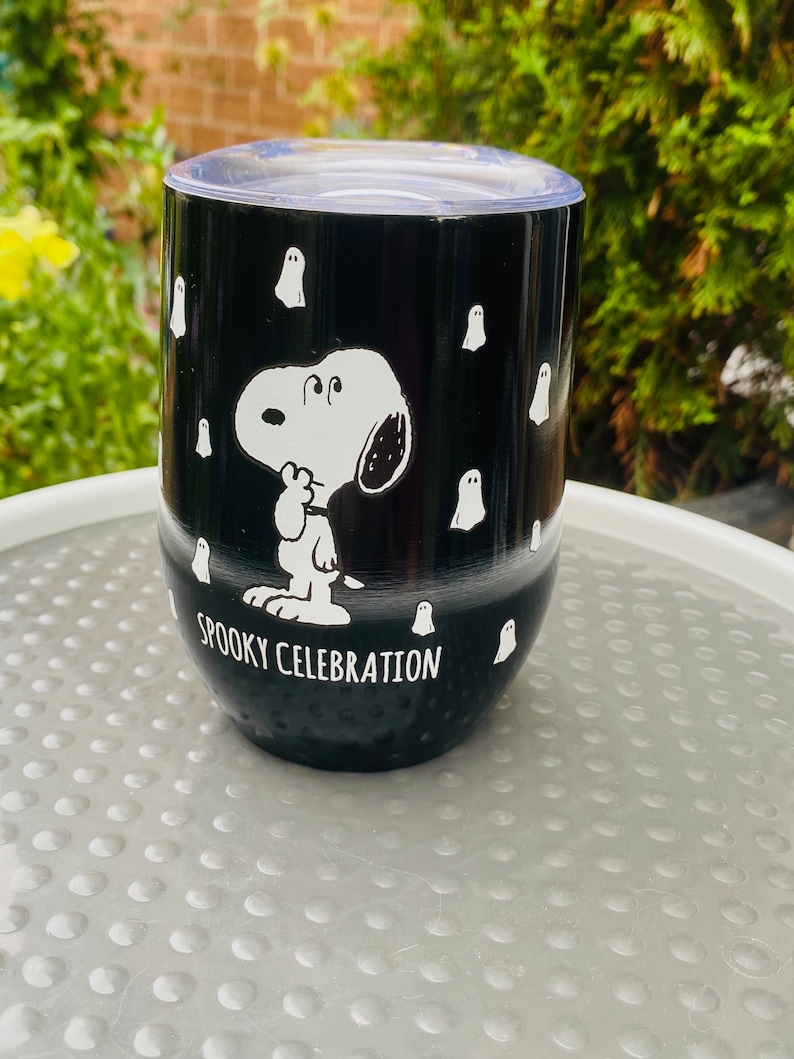 May include: Black stainless steel tumbler with a clear lid. The tumbler features a white illustration of Snoopy and ghosts, with the words "Spooky Celebration". The tumbler is designed for Halloween.