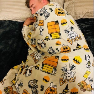 May include: A cream-coloured blanket featuring Snoopy, Charlie Brown, pumpkins, and the phrase "Boo!" in orange and black. The blanket has a Halloween theme and is decorated with cartoon illustrations.