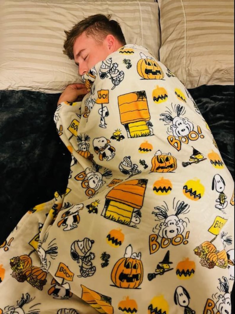 May include: A cream-coloured blanket featuring Snoopy, Charlie Brown, pumpkins, and the phrase "Boo!" in orange and black. The blanket has a Halloween theme and is decorated with cartoon illustrations.