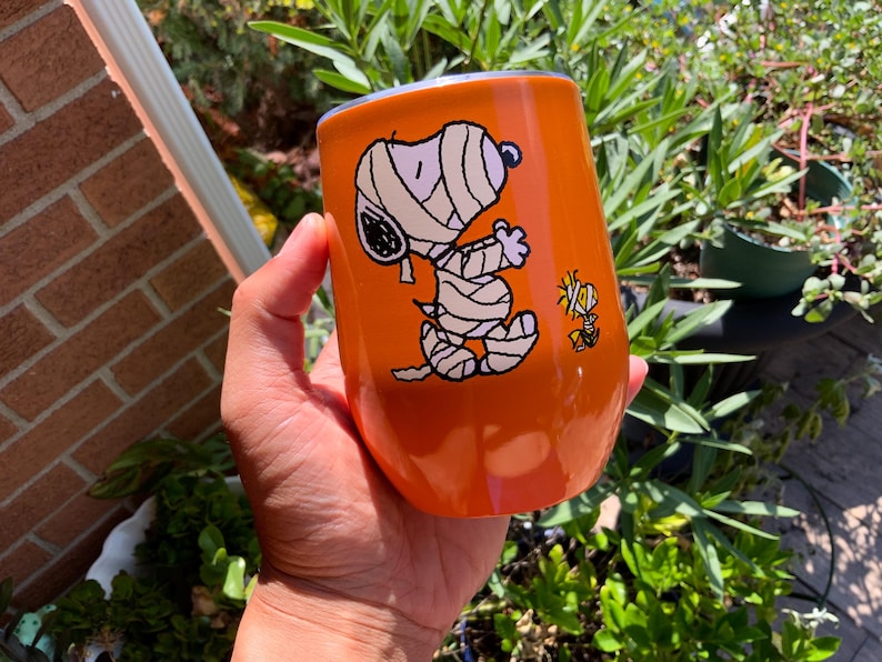 May include: Orange insulated tumbler featuring a Halloween-themed Snoopy design, wrapped in bandages as a mummy, with Woodstock. The tumbler is held in a hand and is surrounded by greenery.