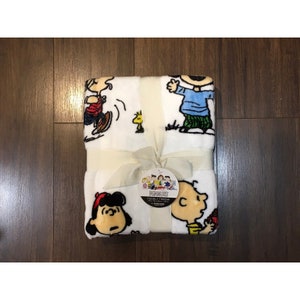 Könnte beinhalten: Eine weiße Peanuts-Decke mit einem Peanuts-Charakterdesign. Die Decke ist mit einem weißen Band gebunden und hat ein Etikett mit der Aufschrift "Peanuts Cuddly Throw".