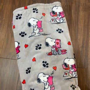 Könnte beinhalten: Eine graue Fleece-Decke mit einem sich wiederholenden Muster von Snoopy, der Valentinstagskarten hält. Die Decke hat rosa Herzen und Pfotenabdrücke.