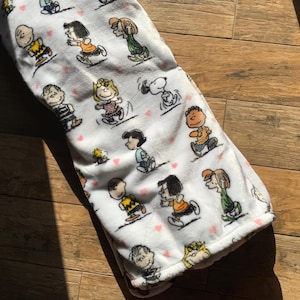 Könnte beinhalten: Weiße Fleece-Decke mit einem sich wiederholenden Muster von Comicfiguren aus den Peanuts. Die Figuren sind alle in verschiedenen Posen und Gesichtsausdrücken, und die Decke ist mit kleinen rosa Herzen verziert.