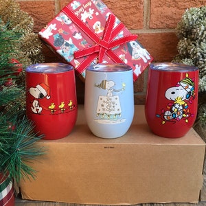May include: Three stemless wine tumblers with holiday-themed Snoopy and Woodstock designs. One red tumbler features Snoopy in a Santa hat. The second is light blue with the text "Be Merry & Bright". The third is red with Snoopy and Woodstock with Christmas lights.