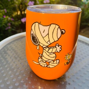 May include: Orange insulated tumbler featuring a cartoon mummy Snoopy and Woodstock. The tumbler has a clear lid and is set against a blurred background of greenery. A fun, themed drinkware item.