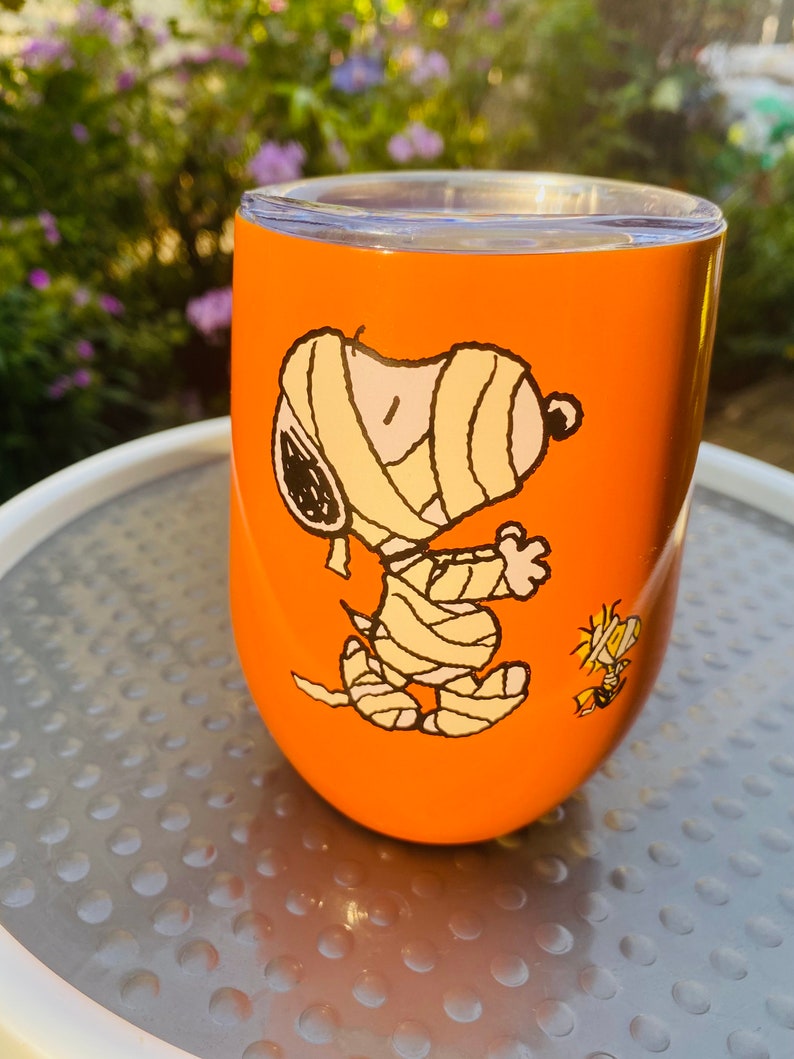 May include: Orange insulated tumbler featuring a cartoon mummy Snoopy and Woodstock. The tumbler has a clear lid and is set against a blurred background of greenery. A fun, themed drinkware item.