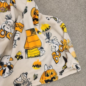 May include: Cream fleece blanket with Halloween-themed Peanuts characters, including Snoopy. The blanket displays pumpkins, the word "BOO!", and autumnal leaves. The soft blanket is folded, revealing its texture.
