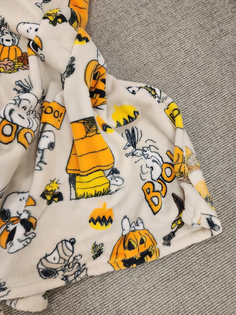 May include: Cream fleece blanket with Halloween-themed Peanuts characters, including Snoopy. The blanket displays pumpkins, the word "BOO!", and autumnal leaves. The soft blanket is folded, revealing its texture.