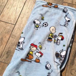 Peut inclure: Une couverture en polaire bleue avec un motif répété de personnages de Snoopy jouant au baseball et à d'autres sports. La couverture présente un fond bleu clair avec des contours blancs des personnages.