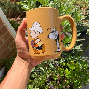 May include: A yellow ceramic mug with a Peanuts cartoon design featuring Charlie Brown and Snoopy. Charlie Brown is wearing a cowboy hat and carrying a book. Snoopy is wearing a red collar and is looking to the left.