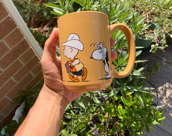 Puede incluir: Una taza de cerámica amarilla con un diseño de dibujos animados de Peanuts con Charlie Brown y Snoopy. Charlie Brown lleva un sombrero de vaquero y lleva un libro. Snoopy lleva un collar rojo y mira hacia la izquierda.