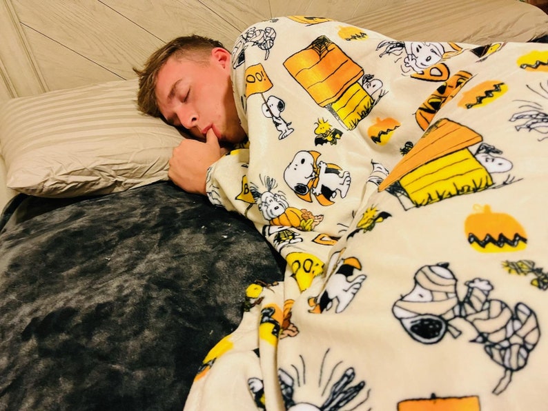 May include: A person asleep under a cream-coloured blanket featuring Snoopy, Woodstock, pumpkins, and Halloween designs. The blanket covers a bed with a grey pillow and a grey blanket, creating a cosy scene.