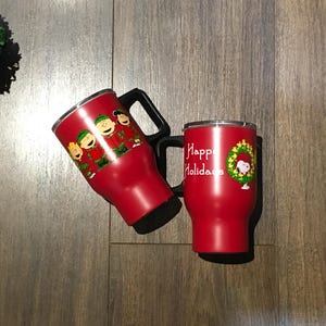May include: Two red insulated tumblers with Christmas-themed designs. One features a group of cartoon characters in winter clothing, while the other says "Happy Holidays" with a wreath and a cartoon character.