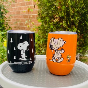 May include: Two stemless wine tumblers featuring Snoopy from Peanuts. One is black with white ghosts and the text "Spooky Celebration." The other is orange with Snoopy as a mummy and Woodstock. Both have silver lids.