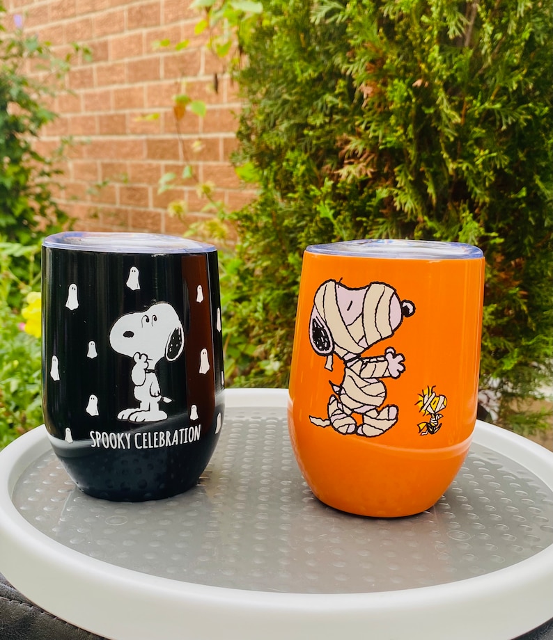 May include: Two stemless wine tumblers featuring Snoopy from Peanuts. One is black with white ghosts and the text "Spooky Celebration." The other is orange with Snoopy as a mummy and Woodstock. Both have silver lids.