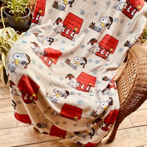 May include: A cream blanket with a repeating pattern of Snoopy in a Santa hat, his red doghouse, snowflakes, and wrapped gifts. The blanket is draped over a wicker chair, creating a cosy, festive feel.