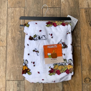 May include: A white plush pet throw blanket featuring a repeating Peanuts pattern with Snoopy, pumpkins, and autumn leaves. A rectangular orange product tag is attached to the centre, displaying the text Peanuts Pet Throw and 127cm x 152cm.