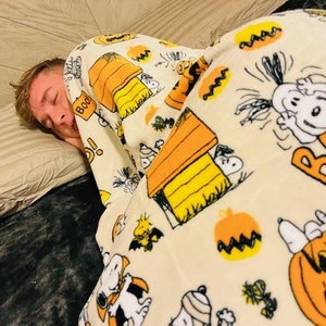 May include: A person asleep under a cream-coloured blanket adorned with a Halloween-themed Snoopy print. The blanket displays Snoopy, pumpkins, and the word "Boo!" in orange. The person is resting on a striped pillow.