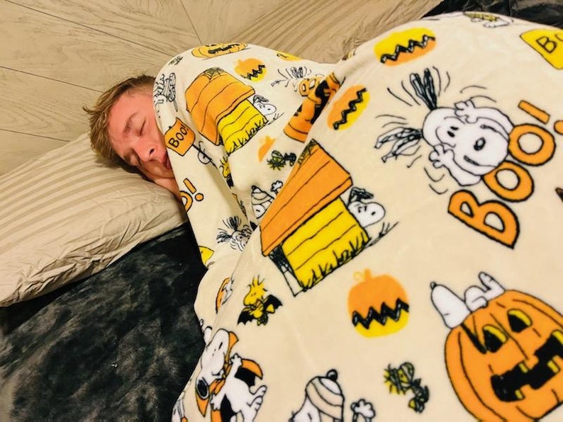 May include: A person asleep under a cream-coloured blanket adorned with a Halloween-themed Snoopy print. The blanket displays Snoopy, pumpkins, and the word "Boo!" in orange. The person is resting on a striped pillow.
