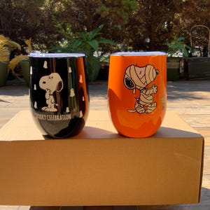 May include: Two stemless wine tumblers with Snoopy designs. One is black with white Snoopy and the text "Spooky Celebration". The other is orange with Snoopy wrapped as a mummy. Both have silver lids and are on a cardboard box.