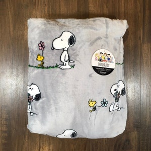 May include: A gray throw blanket featuring Snoopy and Woodstock characters. The blanket has multiple Snoopy and Woodstock illustrations with flowers. The blanket has a tag that reads "Peanuts VelvetSoft Throw".