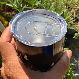 May include: A black tumbler with a clear lid, held in a hand. The tumbler features a repeating pattern of small white ghost illustrations. The lid has a sliding closure. The background includes greenery and sunlight.