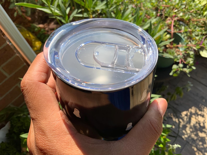 May include: A black tumbler with a clear lid, held in a hand. The tumbler features a repeating pattern of small white ghost illustrations. The lid has a sliding closure. The background includes greenery and sunlight.