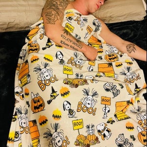 May include: A cream-coloured Halloween-themed blanket featuring Snoopy, pumpkins, and the word "BOO!" in orange and black. The blanket has a repeating pattern of cartoon characters and festive elements.