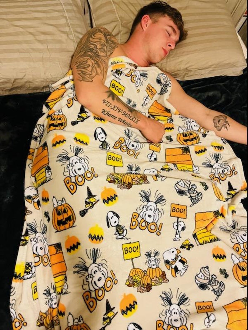 May include: A cream-coloured Halloween-themed blanket featuring Snoopy, pumpkins, and the word "BOO!" in orange and black. The blanket has a repeating pattern of cartoon characters and festive elements.