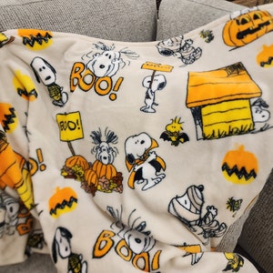 May include: A cream-coloured Halloween-themed blanket featuring Snoopy and Woodstock characters. The blanket has orange pumpkins, the word "BOO!" in orange, and Snoopy in a Halloween costume. The blanket is folded over a grey sofa.