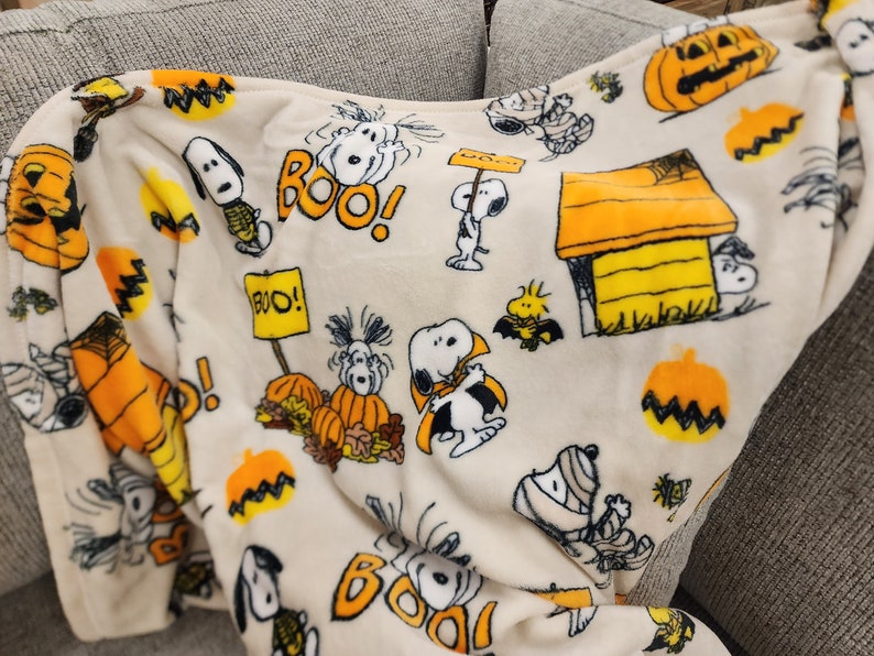 May include: A cream-coloured Halloween-themed blanket featuring Snoopy and Woodstock characters. The blanket has orange pumpkins, the word "BOO!" in orange, and Snoopy in a Halloween costume. The blanket is folded over a grey sofa.