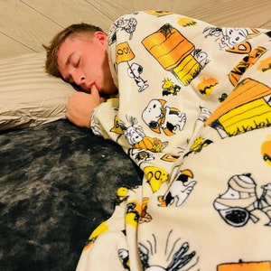 May include: A person asleep under a cream-coloured blanket featuring Snoopy, Woodstock, pumpkins, and Halloween designs. The blanket covers a bed with a grey pillow and a grey blanket, creating a cosy scene.