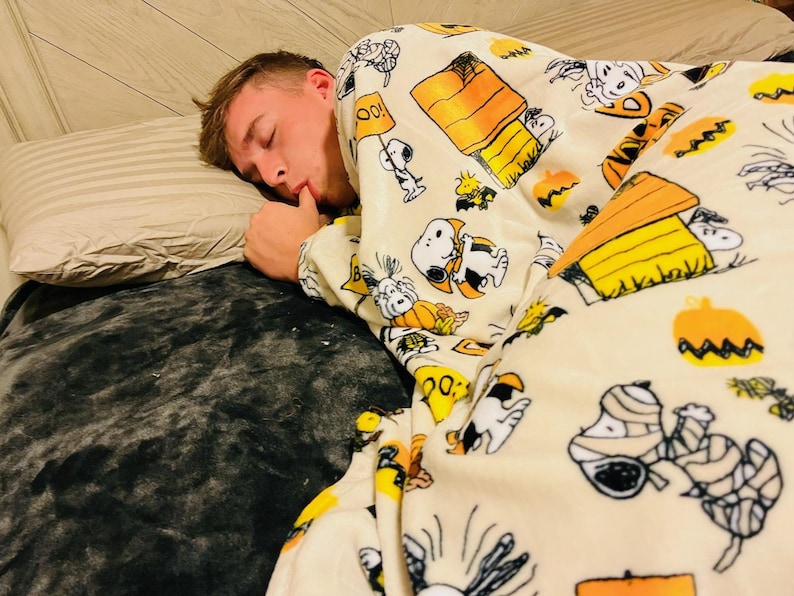 May include: A person asleep under a cream-coloured blanket featuring Snoopy, Woodstock, pumpkins, and Halloween designs. The blanket covers a bed with a grey pillow and a grey blanket, creating a cosy scene.