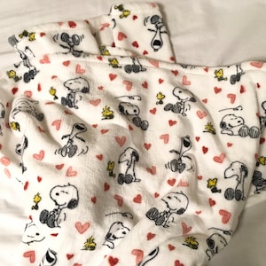 May include: White fleece blanket featuring a repeating pattern of Snoopy and Woodstock. Snoopy is shown in various poses, including sitting, standing, and dancing. Woodstock is depicted flying and perched on Snoopy's head. The blanket is decorated with small pink hearts.